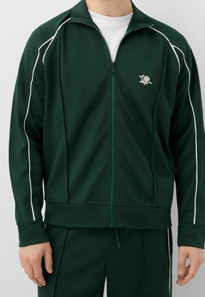 Mężczyzna nosi ciemnozieloną kurtkę dresową z białym obszyciem na rękawach i małym białym emblematem róży na piersi, w połączeniu z pasującymi spodniami.