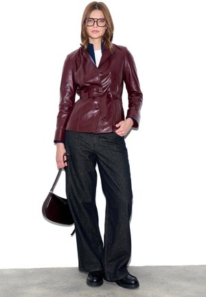 Kvinde stående iført en vinrød læderjakke med bælte, sorte vidde bukser, sorte sko, store briller og med en mørkebrun håndtaske i hånden.