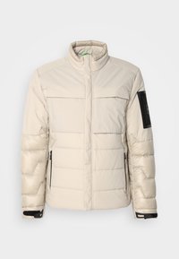 Veste d'hiver matelassée beige avec col montant, deux poches à rabat sur la poitrine, poches latérales zippées et patch noir sur la manche gauche.