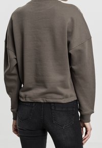 Sudadera marrón de gran tamaño que presenta un cuello redondo acanalado y mangas de corte holgado, con un acabado texturizado y dobladillo elástico. Combinada con jeans oscuros.