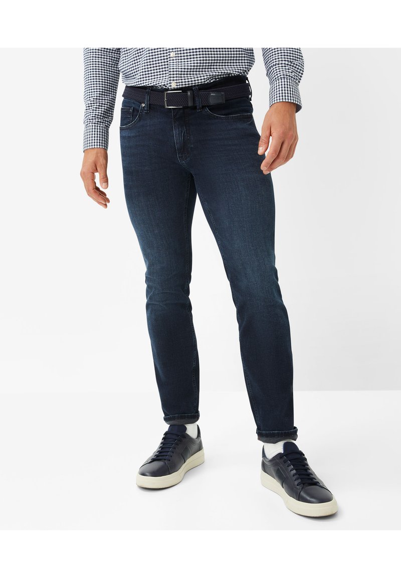 Dark blue jeans with a slim fit and slight stretch, paired with navy sneakers featuring white soles, worn with a checkered shirt.