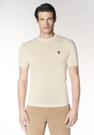 Uomo che indossa un maglione beige aderente a maniche corte con un piccolo logo ricamato sul petto e pantaloni marroni chiari.