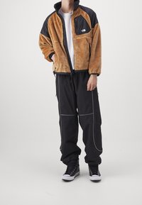 Giacca in pile zip-up marrone e nera con colletto alto, grande tasca frontale e pantaloni neri con cuciture bianche a contrasto. Sneakers nere.