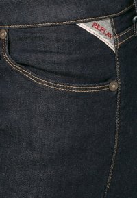Jeans de mezclilla oscura con costuras tonales, herrajes de botón metálico y una etiqueta de logo discreta en rojo en el bolsillo frontal. Textura suave.