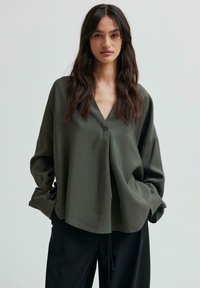 Camicia verde oliva con bottoni, scollatura a V, maniche lunghe, vestibilità over-size e tessuto morbido. Indossata con pantaloni neri.