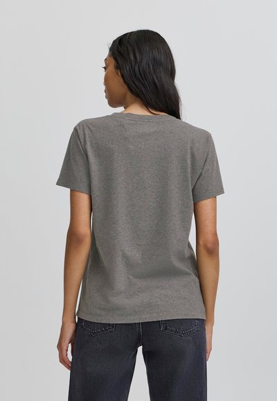 Grå kortærmet T-shirt med rund hals, fremstillet af bomuldsmateriale. Subtil tekstur; parret med mørke jeans i baggrunden.