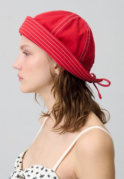Cappello in cotone rosso con cuciture bianche, di forma rotonda, con lacci sul retro. Indossato da un modello con capelli castani di lunghezza media.