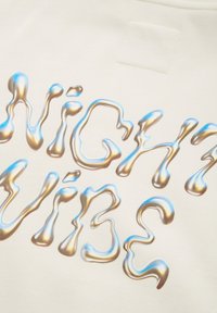 Il tessuto color crema presenta il testo "NIGHT VIBE" in un carattere argento e blu lucido, simile a un liquido, mostrando una texture liscia e riflettente.