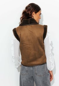 Gilet sans manches marron avec un col doux et texturé. Porté sur une chemise blanche à manches longues à volants, associé à un jean taille haute gris.