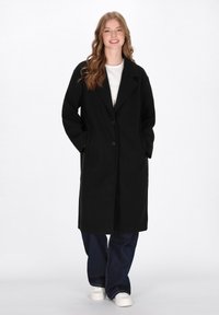 Abrigo de mezcla de lana negra con solapa muesca, cierre de un botón y bolsillos laterales. Llevado sobre una camisa blanca y combinado con jeans oscuros.