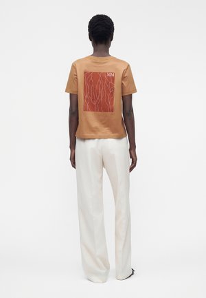 Persoană stând cu fața înapoi, purtând un tricou maro cu un design abstract roșu și pantaloni albi pe un fundal simplu alb.