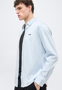 Jeune homme portant une chemise à boutons bleu clair avec le logo "BOSS" sur un t-shirt noir, debout dans une pose décontractée contre un fond uni.