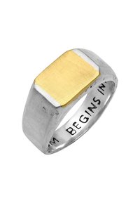 Metallring med en rektangulär gul topp, silver-texturerat band och ingraverad text inuti som säger "BEGINS IN." Geometrisk design med kontrasterande ytor.