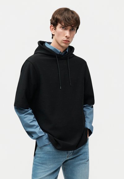 Νέος άνδρας με καστανά μαλλιά, φορούσε μαύρη κοντομάνικη φούτερ με κουκούλα πάνω από μπλε μακρυμάνικο πουκάμισο και ανοιχτόχρωμα τζιν, με τα χέρια στις τσέπες.