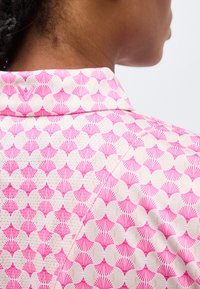 Ο λαιμός και ο ώμος ενός ατόμου που φοράει ενδυμασία μπεζ και ροζ με σχέδια σε σχήμα βεντάλιας και εμφανείς ραφές.