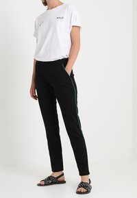 Vit bomull t-shirt med trycket "WAIT", ihop med svarta byxor med en grön sidremsa. Buren med svarta sandaler dekorerade med nitar.