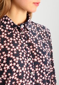 Dunkelblaue Bluse mit einem rot-weißen Sternenmuster, Knopfleiste, Kragen und Taschen mit Detail. Aus leichtem Stoff mit glatter Textur hergestellt.