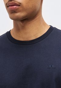 Κοντινό πλάνο ενός άνδρα που φοράει μπλε σκούρο μπλουζάκι με στρογγυλή λαιμόκοψη και μικρό ανάγλυφο λογότυπο στο αριστερό μέρος του στήθους, με φόντο μια μονή απόχρωση.