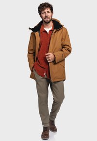 Brown waterproof jacket with a black collar, worn over a red corduroy shirt, paired with beige pants and brown lace-up boots.