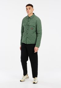 Groene gewatteerde jas met een knoopsluiting, twee borstzakken en zwarte accenten, gecombineerd met zwarte broek en lichtgekleurde schoenen.