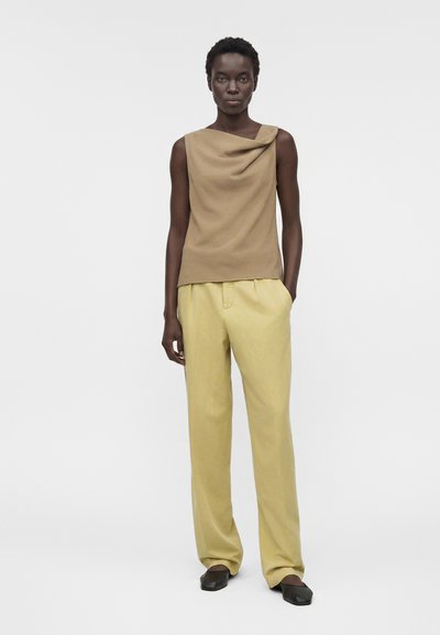 Μαύρη γυναίκα στέκεται με τα χέρια στις τσέπες, φοράει αμάνικη μπεζ μπλούζα, χαλαρό ανοιχτό κίτρινο παντελόνι και μαύρα σανδάλια με επίπεδο τακούνι σε λευκό φόντο.