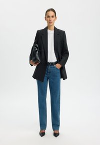 Zwarte blazer over een wit T-shirt, gecombineerd met lichtblauwe high-waisted jeans. Houdt een zwarte handtas vast, met puntige zwarte hakken.
