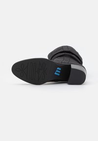 Botín negro con parte superior texturizada, tacón bajo y suela de goma que presenta un logo azul y un patrón decorativo. La vista inferior muestra un diseño de banda de rodadura detallado.