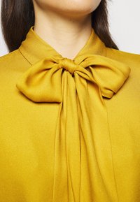 Gros plan sur un chemisier en satin jaune moutarde avec un grand nœud noué au col, porté par une personne aux cheveux bruns.