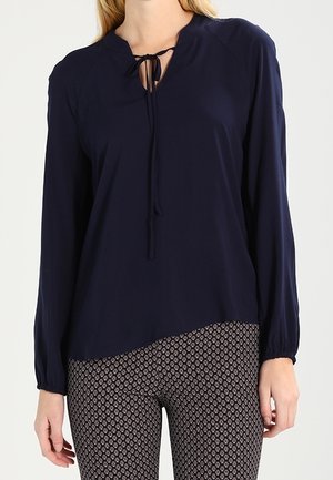 Kobieta ubrana w granatową bluzkę z długim rękawem z wiązaniem przy dekolcie oraz dopasowane spodnie w wzory na jednolitym tle.