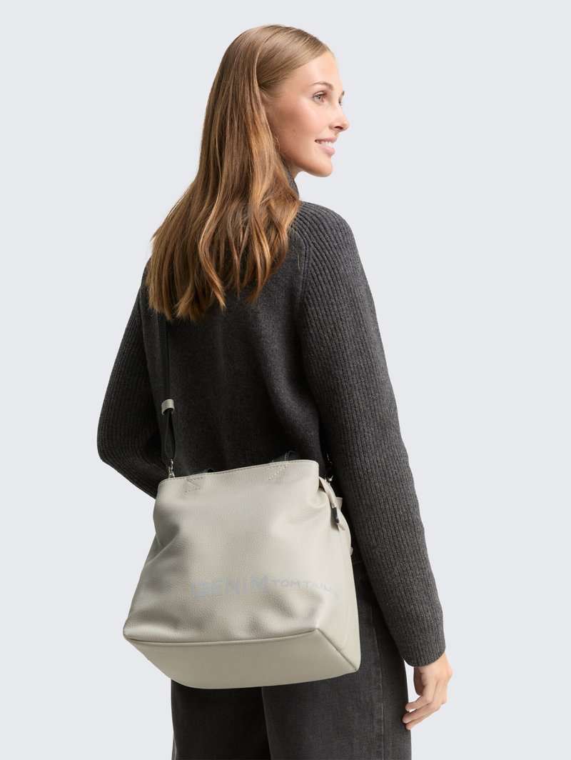 Woman with long light brown hair wearing a dark gray sweater and carrying a light gray shoulder bag with thin black strap.
