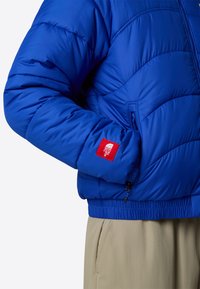 Giacca puffer blu con texture trapuntata, polsini e orlo elasticizzati. Presenta una toppa rossa con logo sulla manica. Abbinata a pantaloni beige.