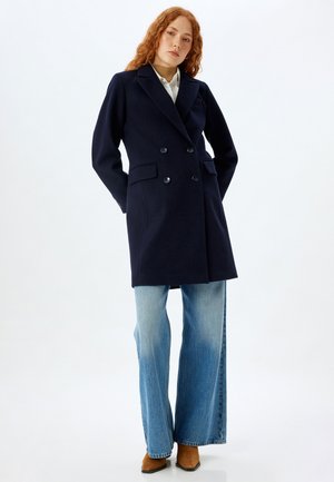 Navy double-breasted coat with two front pockets, notched lapels, and a smooth texture, paired with wide-leg light blue jeans and brown shoes.