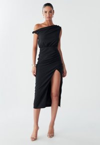 Model stands confidently in a sleek off-shoulder black dress with a thigh-high slit, accessorized with gold jewelry and light heels.