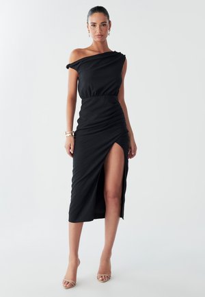 Model staat zelfverzekerd in een elegant zwart off-shoulder cocktailjurk met een hoge split, geaccessoireerd met gouden sieraden en lichte hakken.