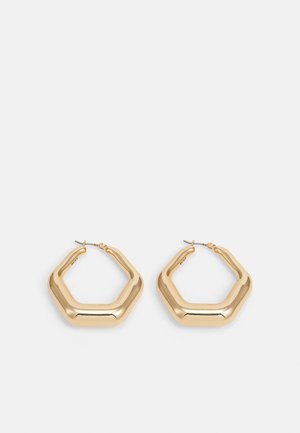 Paire de boucles d'oreilles créoles hexagonales épaisses dorées avec surfaces lisses et polies et fermoirs à poussette sur fond blanc.