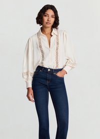 Blouse en coton crème avec des détails en dentelle sur le devant, manches bouffantes et fermeture à boutons, associée à un jean ajusté bleu foncé.