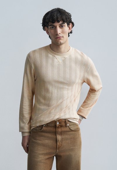Jersey de manga larga beige claro con patrón espiga, cuello redondo y puños acanalados, combinado con jeans de mezclilla marrón claro.