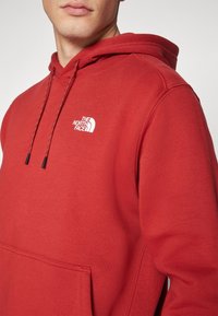 Rote Kapuzenjacke aus weichem Material mit einer Kängurutasche. Verfügt über eine verstellbare Kapuze mit Kordelzug und ein weißes Logo auf der Brust.