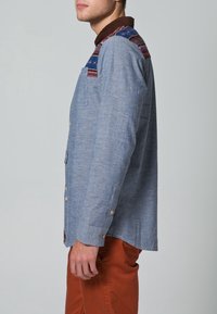 Camisa azul claro con cuello marrón y detalles de colores en los hombros; cuenta con un bolsillo delantero y puños con botones.