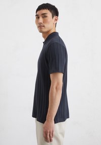 Marineblauwe geribde polo met korte mouwen en een kraag, met subtiele verticale lijnen en een getailleerd ontwerp. Glad textuur, minimale hardware.