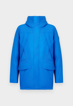 Mėlyna striukė su aukštu apykakle, dviem didelėmis priekinėmis kišenėmis ir elegantišku, minimalistiniu dizainu su ryškiomis siūlės detalėmis.