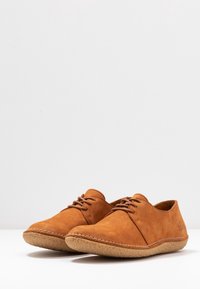 Une paire de chaussures décontractées en suède marron à lacets avec des semelles en caoutchouc texturé marron clair, sur un fond et une surface blanche.