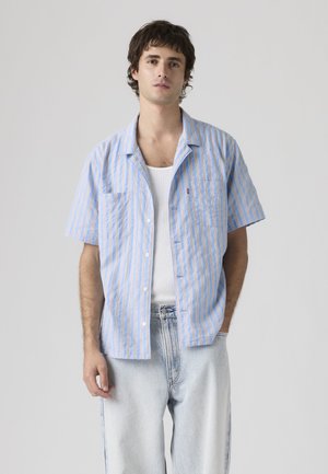 Jeune homme portant une chemise à manches courtes rayée bleu clair et beige sur un débardeur blanc et un jean bleu clair, debout devant un fond uni.
