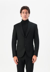 Black tailored suit with a notch lapel, two-button closure, and a slim fit. Worn over a black turtleneck sweater, minimalistic design.