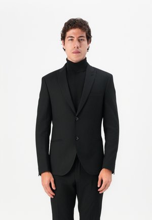 Traje negro a medida con solapa de muesca, cierre de dos botones y corte slim. Llevado sobre un suéter negro de cuello alto, diseño minimalista.