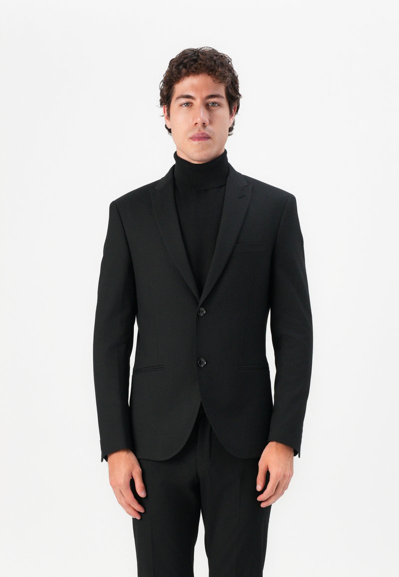 Black tailored suit with a notch lapel, two-button closure, and a slim fit. Worn over a black turtleneck sweater, minimalistic design.