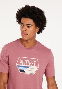 Rožinė medvilnė marškinė su grafiniu „PROTEST“ logotipu, kuriame yra horizontalių juostų mėlynos ir grietinėlės spalvų. Trumpomis rankovėmis ir apvaliu iškirpimu.