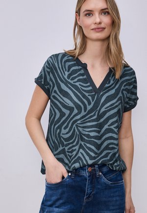 Kobieta ubrana w krótki top w paski zebry z dekoltem w kształcie litery V, wsunięty w niebieskie dżinsy, z jedną ręką w kieszeni, na jednolitym tle.