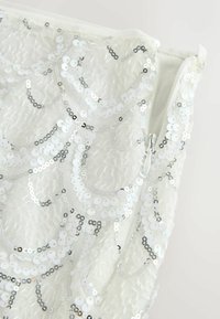 Tissu blanc avec des sequins irisés en motifs coquilles et une fermeture éclair blanche partiellement ouverte sur le côté droit.