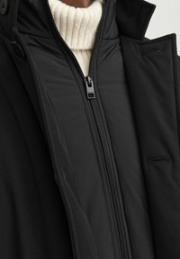 Jachetă neagră cu fermoar în față, având un guler înalt crem vizibil dedesubt. Material texturat și detalii de design ajustate.